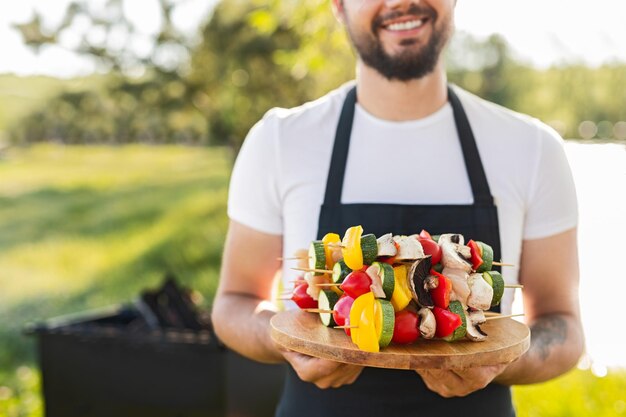 Crop man holding tray with raw veggie kebabs before cooking in nature