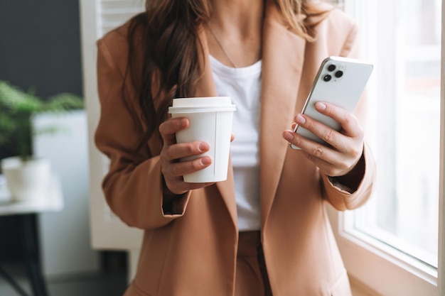 現代のオフィスで手にコーヒーとスマートフォンで紙コップを保持しているベージュのスーツのビジネス女性の作物の写真