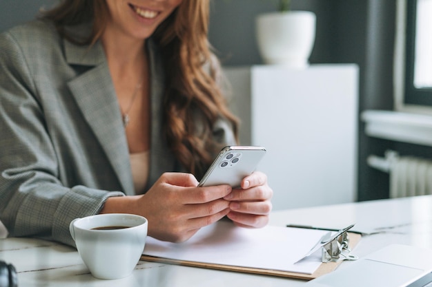 明るい現代のオフィスの女性で働く手で携帯電話を使用してスタイリッシュなスーツで長い髪の若い笑顔のブルネットの女性の作物の写真はスマートフォンでメッセージを送信します