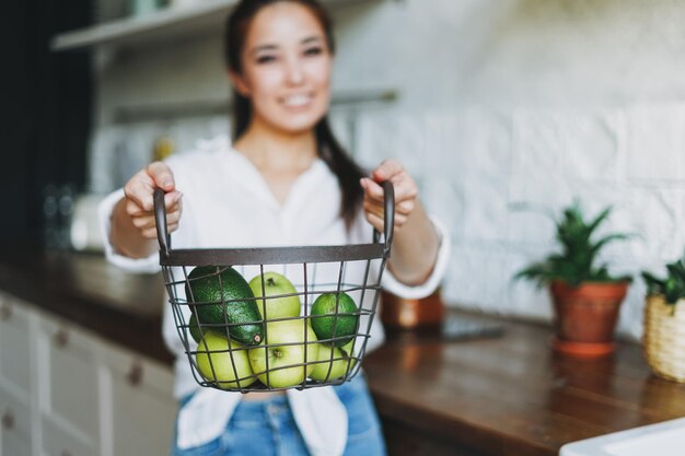 Crop photo of young woman in white shirt hold basket with green fruits and vegetables in hands on the kitchen