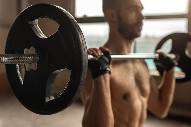 Crop shirtless male athlete in hand wraps looking forward while raising barbell during workout in gymnasium