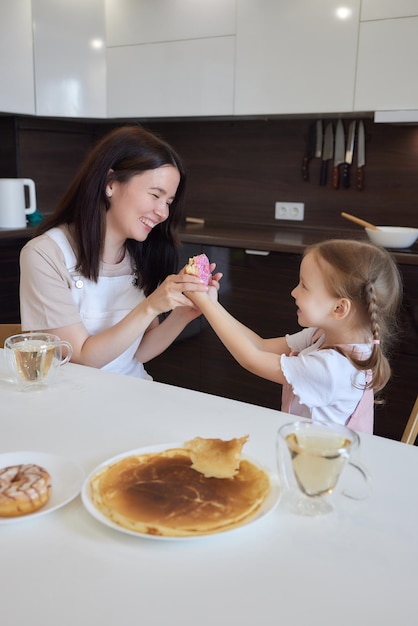 Ritaglia la madre in rosso e sua figlia con ciambelle colorate sedute in cucina concetto di dieta e cibo spazzatura