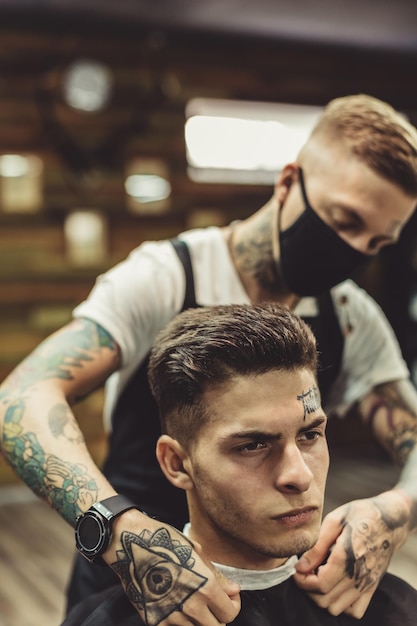 Crop stylist preparing young tattooed man in chair for shaving in barbershop