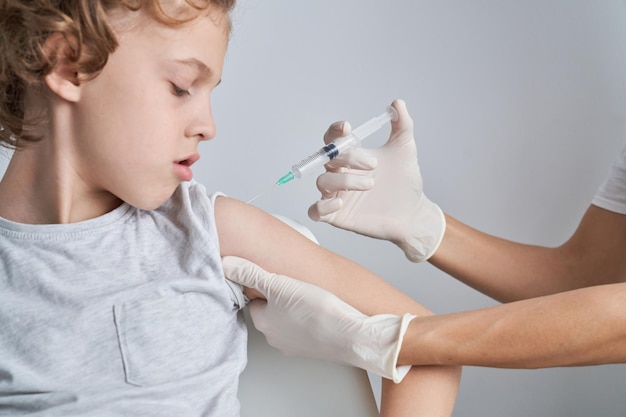 Crop unrecognizable doctor in latex gloves injecting vaccine into shoulder of boy on white background during epidemic in light room