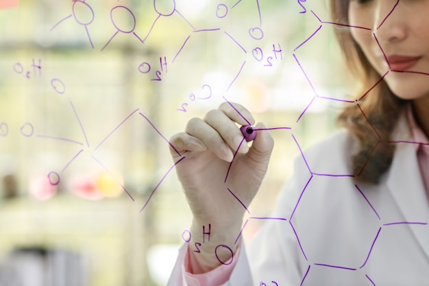 Crop unrecognizable female scientist with marker writing chemical formulas on glass board while working with colleagues in laboratory.