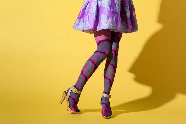 Crop unrecognizable female with sexy legs wearing fashionable purple ornamental collant with mini skirt and high heeled shoes on yellow background with shadow