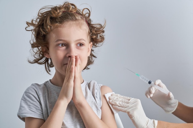 Crop unrecognizable medic with syringe wiping shoulder of cute boy with folded hands while preparing for injecting in white background