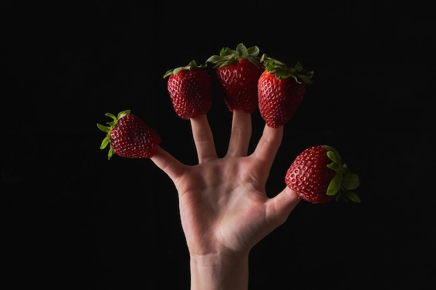 Crop unrecognizable person wearing appetizing red strawberries on fingers on black background in studio