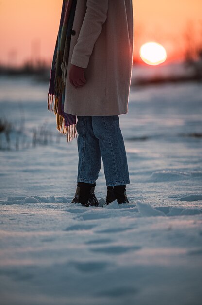 Crop unrecognizable woman in warm clothes and boots on fresh white snow in winter countryside at sunset time