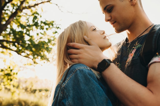 Crop view of young beautiful couple looking each other in eye and hugging, man touching hair ofï¿½attractive young blondï¿½woman standing outdoors on blurred backlit background