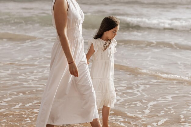 crop woman holding hand of adorable little daughter while strolling on seashore