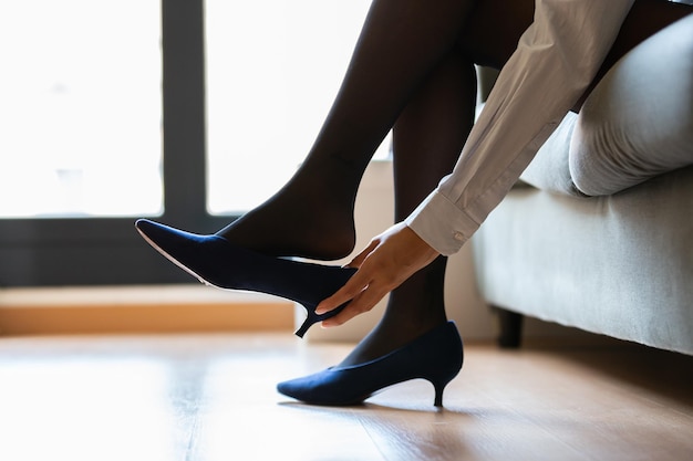 Crop young woman taking off her heels at home due to pain and relaxing on the sofa