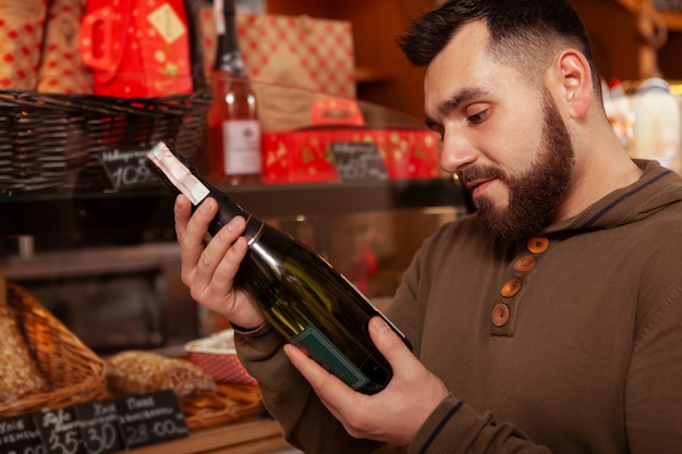 Обрезанный крупным планом бородатый мужчина изучает бутылку вина, делая покупки для празднования праздников