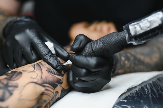 Cropped close up of a bearded tattoo artist working at his studio tattooing sleeve on the arm of his male client. Man getting tattooed by professional tattooist