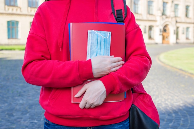 Фото Обрезанное фото умного умного подростка в повседневном красном джемпере с блокнотами и медицинской маской в руках крупным планом
