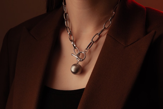 Cropped close up portrait of young woman with perfect silky skin, demonstrating silver jewelry chain on the neck in a brown jacket.