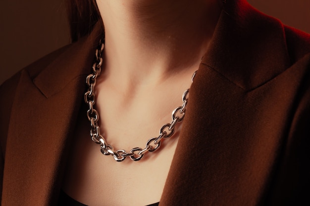 Cropped close up portrait of young woman with perfect silky skin, demonstrating silver jewelry chain on the neck in a brown jacket.