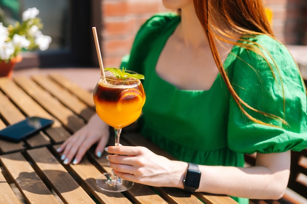 Cropped close-up shot of unrecognizable young woman holding glass with cold exotic cocktail sitting at table in outdoor cafe in sunny summer day. Pretty female hipster student drinking cool lemonade.