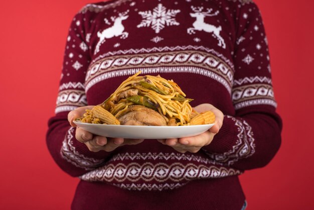 赤と白のクリスマス セーターを着た女の子のトリミングされたクローズ アップ写真は、孤立した赤の背景に脂肪分の多い食品の大皿を保持しています。