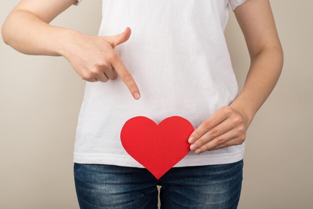 Cropped closeup photo of girl in white tshirt and jeans points her finger at red paper heart near crotch on isolated grey background