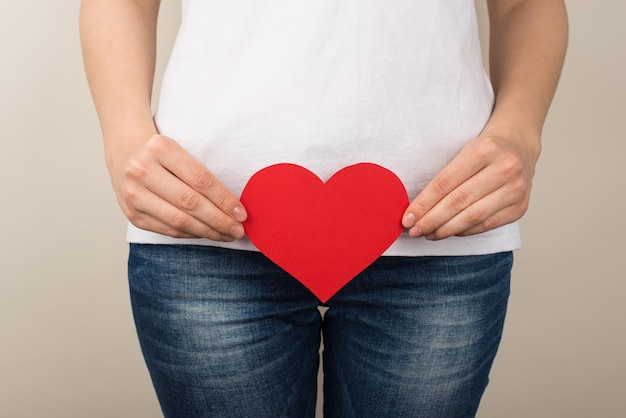 Cropped closeup photo of woman in white tshirt and jeans holding red paper heart near crotch on isolated grey background