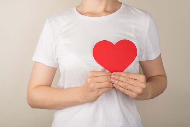 Cropped closeup photo of young woman in white tshirt holding red paper heart near heart on isolated grey background