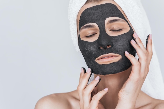 Cropped closeup portrait of beautiful young woman applying clay black mask doing beauty wellness treatment on her face skin Horizontal shot of female applying organic facial mask with closed eyes