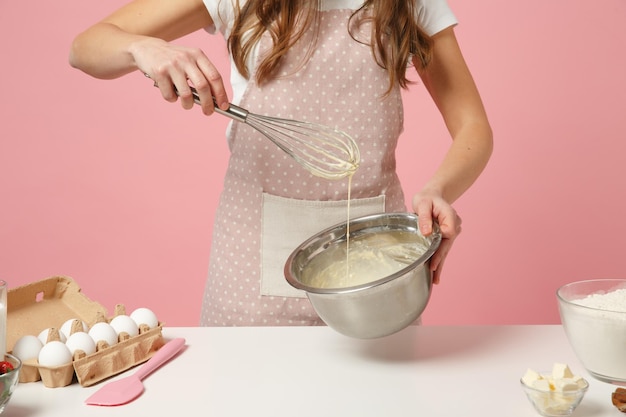 Cropped female chef cook confectioner or baker in apron white t-shirt cooking cake or cupcake at table hold whisk bowl isolated on pink pastel background in studio. Mock up copy space food concept.