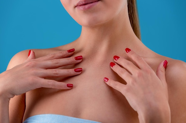 Cropped frontview of model touching her face and neck tenderly. Feminine hands with accurete nails, pink plump lips. Posing on blue studio background. Having smooth, healthy skin tone