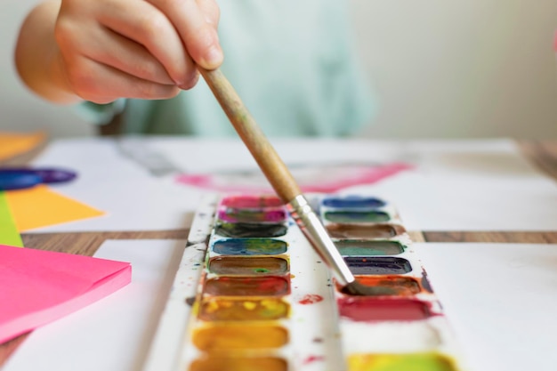 教室で絵を描きながらパレットの上にペイントブラシを握っている男の子のカットされた手