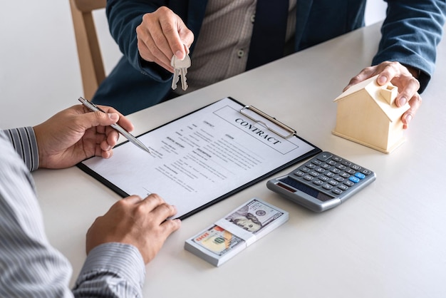 Photo cropped hand of client signing document while businessman holding key at table in office
