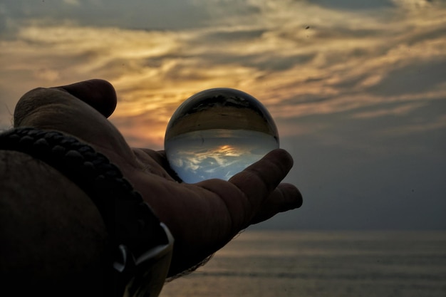 Foto mano tagliata che tiene una palla di cristallo contro il mare durante il tramonto