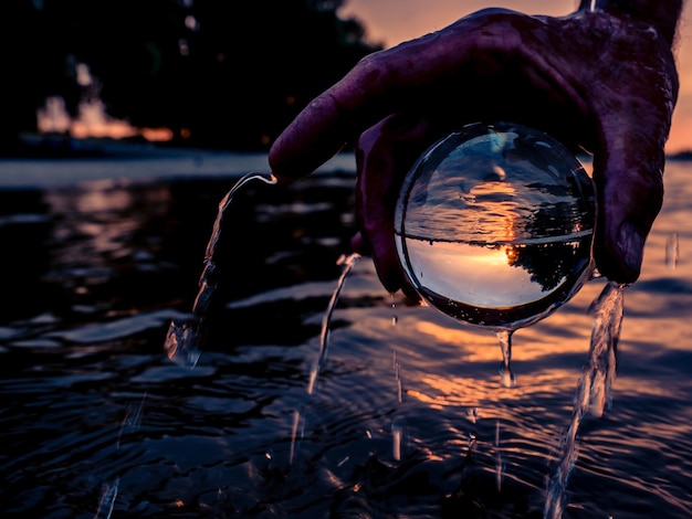 Foto mano tagliata che tiene una palla di cristallo in mare durante il tramonto