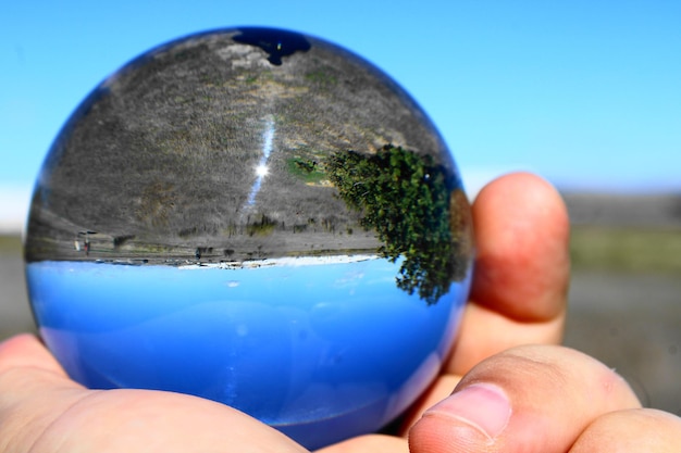 Foto mano tagliata che tiene una palla di cristallo con il riflesso del paesaggio contro un cielo limpido