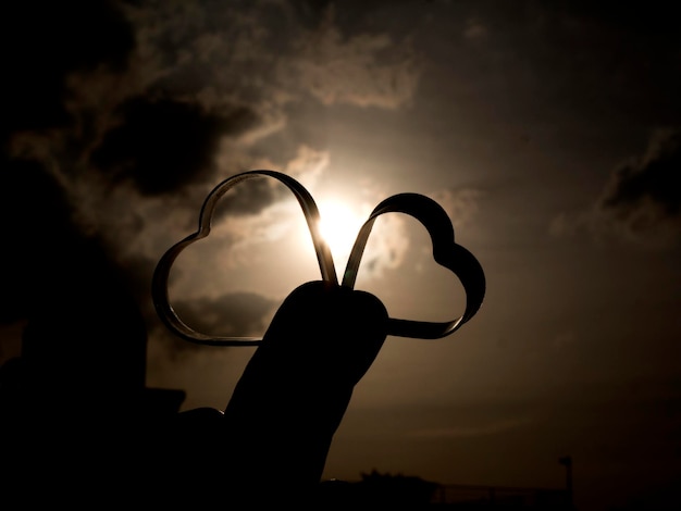 Foto mano tagliata che tiene una forma di cuore contro il cielo durante il tramonto