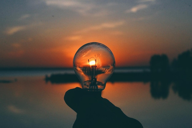 Foto mano tagliata che tiene una lampadina vicino al lago contro il cielo durante il tramonto