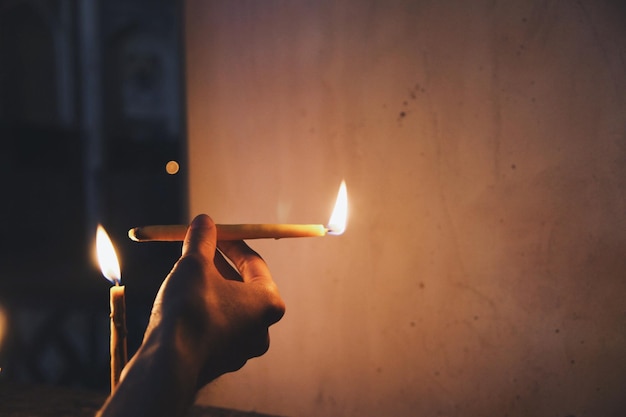 Cropped hand holding lit candle against wall