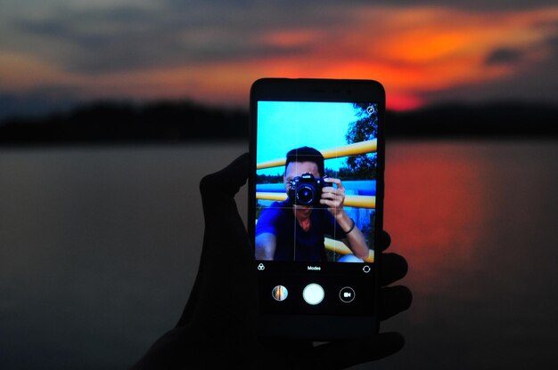 Foto mano tagliata che tiene il telefono cellulare al lago contro un cielo nuvoloso durante il tramonto