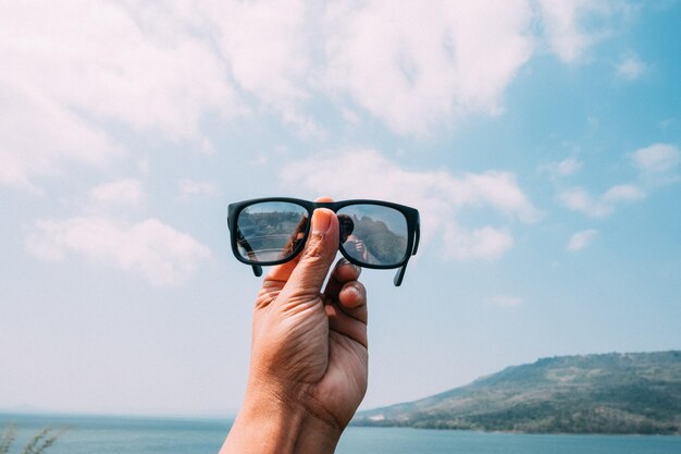 Foto mano tagliata che tiene gli occhiali da sole contro il mare