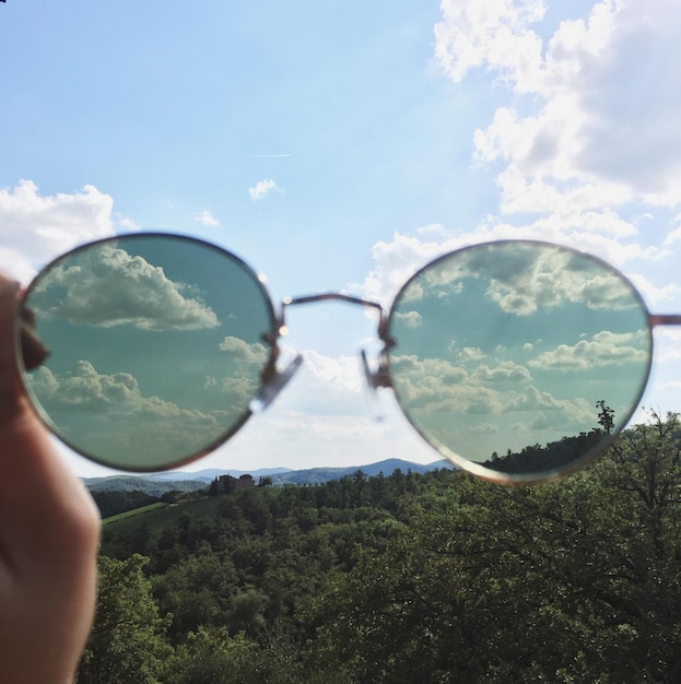 Foto mano tagliata che tiene gli occhiali da sole contro il cielo