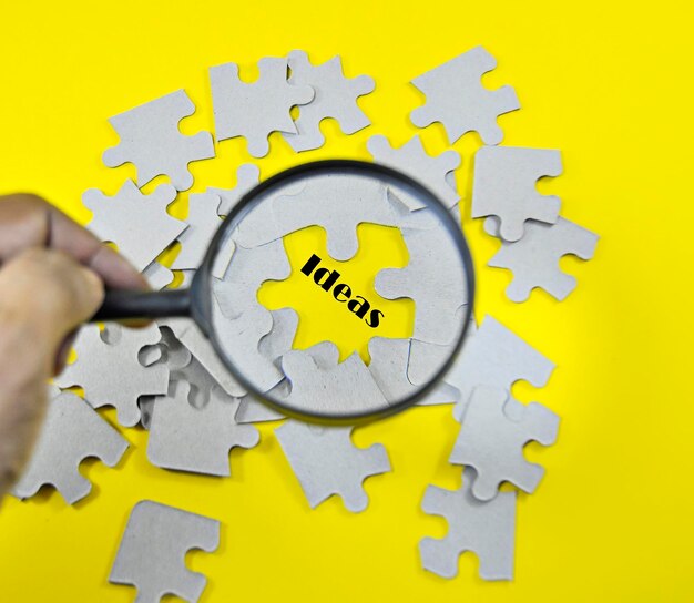 Cropped hand looking ideas text with magnifying glass by jigsaw pieces on yellow background