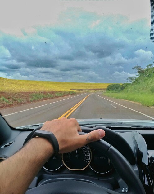 Cropped hand of man driving car on road against sky