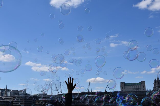 Cropped hand of person holding bubbles against blue sky