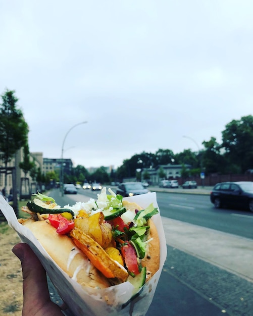 Foto mano tagliata di una persona che tiene il cibo per strada in città contro il cielo