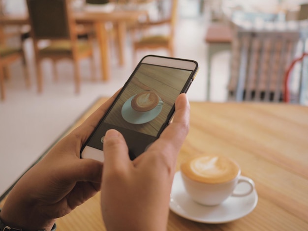 Cropped hand of person photographing coffee through mobile phone on table