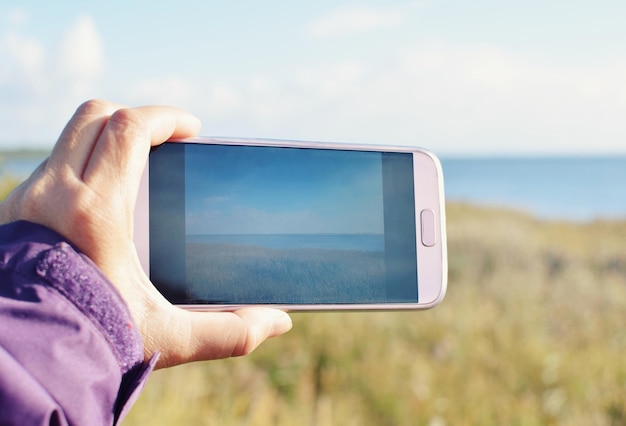 Cropped hand photographing sea against sky through mobile phone