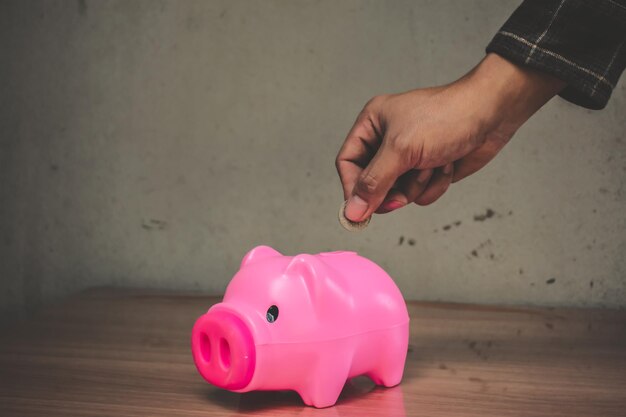 Cropped hand putting coin in piggy bank on wooden table against wall