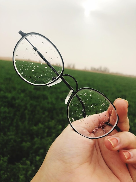 Photo cropped hand of woman holding wet eyeglasses against sky