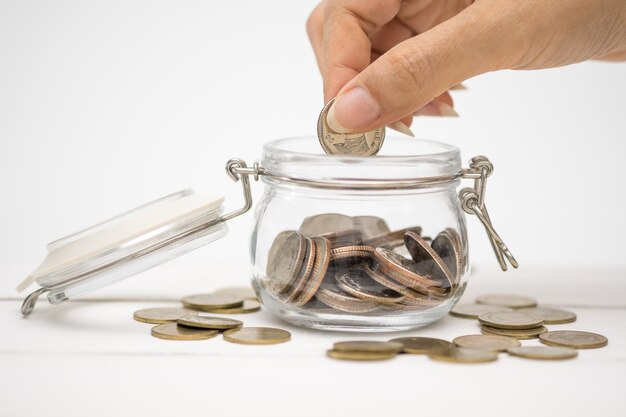 cropped hand of woman putting coins into glass jar against white background
