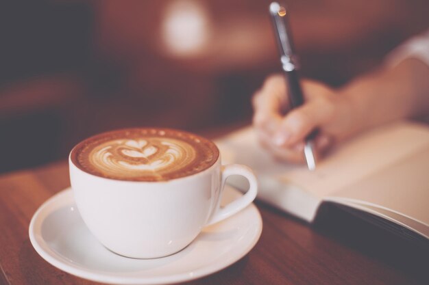Cropped hand of woman writing on book by coffee on table